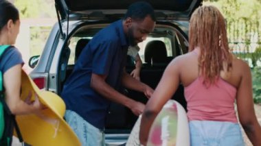 Diverse couple with mother and daughter putting baggage inside vehicle while going on holiday summer citybreak. Diverse people loading car trunk with voyage luggage while kid hurrying them