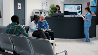 Afrikalı Amerikalı doktor emekli bir hastayla tedaviyi tartışıyor, hastane resepsiyonunda sağlık hizmetleri teşhisinden bahsediyor. Bekleme odasında çok ırklı insanlar sağlık kontrolü yapıyor..