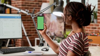 Female employee holding smartphone with greenscreen display at home, working remotely. Woman looking at mobile phone with isolated chroma key, blank copyspace template and mockup background.