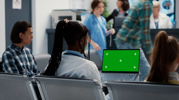 African american physician working with greenscreen on laptop computer, sitting in hospital reception lobby. Medic using isolated chroma key background with blank copyspace and mockup template.
