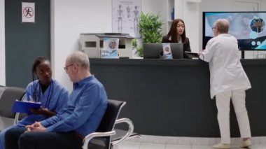 Female receptionist talking to senior doctor at reception counter in medical facility waiting area. General practitioner asking worker about medical reports and healthcare appointments.