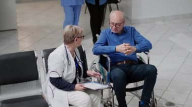 Wheelchair user with caretaker having consultation with medic in waiting area at facility. Old patient with disability receiving nursing assistance and medical checkup from physician. Handheld shot.