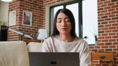 Focused freelancer woman working remotely from home on modern laptop. Beautiful young adult person doing remote freelancing work on portable computer while sitting on sofa inside apartment.