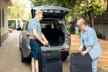 Elderly husband and wife going on holiday vacation and putting luggage or travelling bags in automobile trunk. People leaving on retirement trip journey with suitcases and trolley in vehicle.