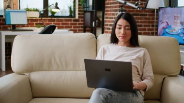 Focused woman sitting on sofa while working remotely on laptop. Beautiful young adult asian student attending online class while typing important lessons on modern portable computer at home.