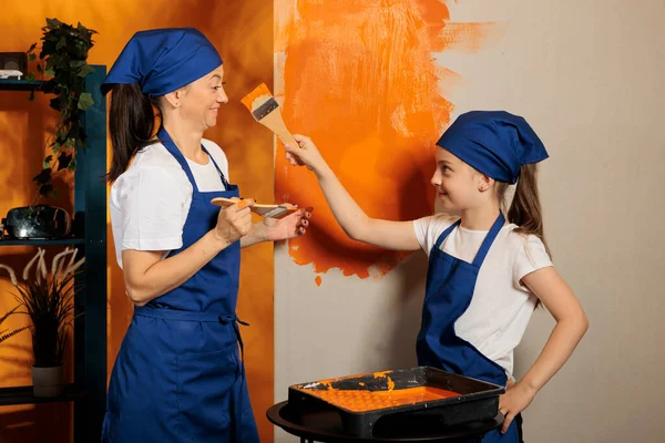 Family having fun with orange paint, using color to renovate apartment walls together. Woman with child laughing and painting house room with paintbrush tools and, diy interior job.