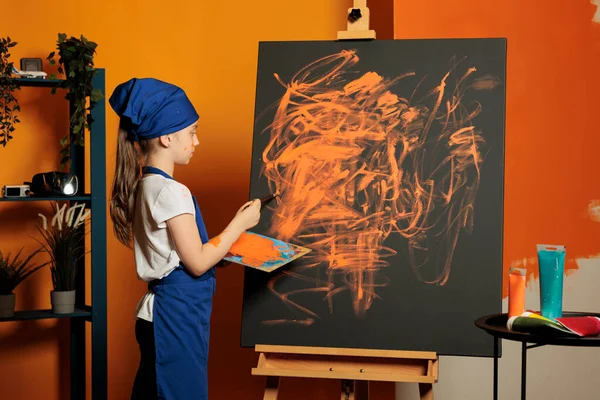 Young child using orange paint color on canvas, creating artwork masterpiece with colormix tray and aquarelle palette. Painting inspiration design with painbrush, watercolor and skills.