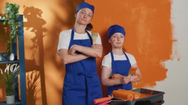 Portrait of family renovating apartment space with tools and orange paint to color house walls. Woman with little girl working on home interior redecoration with paintbrush roller.