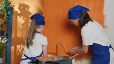 Mother and little kid using roller and orange paint to renovate apartment walls for housework improvement. Painting room interior with paintbrush and color, working on redecoration and renovation.