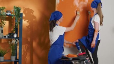 Small family painting apartment walls with orange color, using ladder and paintbrush to work on house interior redecoration. Woman and young kid renovating home with tools. Tripod shot.