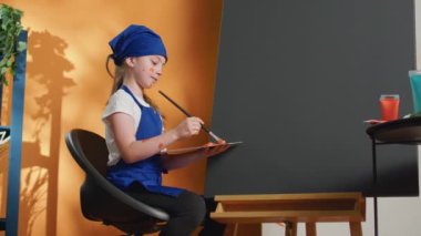 Young kid painting artwork with orange color on canvas, using aquarelle paint with paintbrush and watercolor mixing tray. Creating masterpiece with artistic tools and brushes, creative vision.