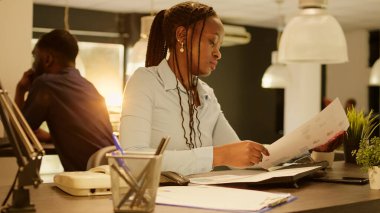 African american woman analyzing business paperwork with charts and data research to work on online project report. Brainstorming ideas to plan network presentation on computer during sunset.