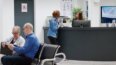 African american patient talking to receptionist about medical consultation, having appointment with medic physician. Woman asking for help at clinic reception counter in waiting area.