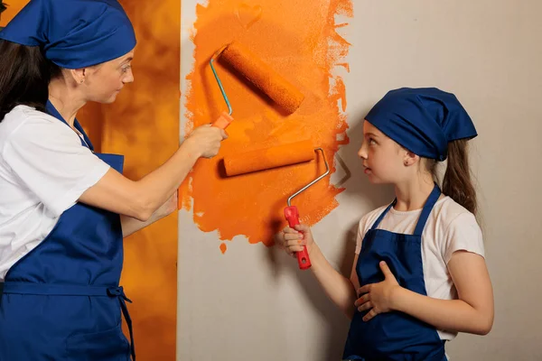 Mother and kid using orange paint on roller painbrush to change wall color, working on house redecoration. People having fun painting apartment dcor interior, renovating with tools and roll.