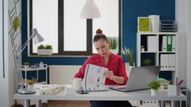 Professional assistant analyzing financial statistics on files and laptop, using marketing report to create charts and graphs. Sales consultant working on business strategy in startup office.