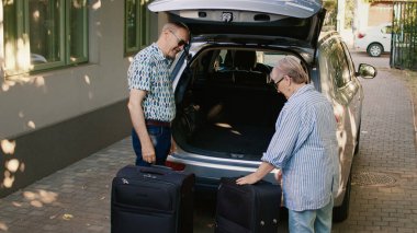 Cheerful elders going on retirement voyage while having heavy baggage and travel trolley. Senior couple putting luggage in car trunk while getting ready for holiday trip.