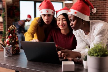 Diverse team of women working on startup business in festive office with christmas tree and decorations. Doing teamwork and celebrating xmas time with seasonal holiday ornaments.