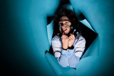 Mad scientist with messy hairstyle and filled glass test tubes grinning dreadful at camera while on blue background. Foolish maniac chemist with having tray with test tubes filled with toxic compounds