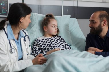 Caring father sitting next to sick daughter resting in hospital pediatric ward while doctor presenting treatment scheme. Pediatrician examining ill kid health condition while discussing with parent.