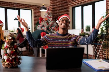 Portrait of african american festive man at office job decorated with christmas tree and seasonal ornaments. Happy employee working on laptop during winter season holiday, xmas decorations.
