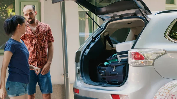 Caucasian family loading voyage luggage inside car trunk while going on summer holiday field trip together. People getting ready for weekend citybreak while putting baggage inside vehicle.