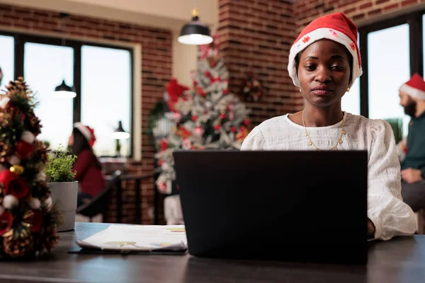 Company worker using laptop at startup office, celebrating winter season with christmas decorations and tree. Woman with santa hat working in space with festive seasonal ornaments.