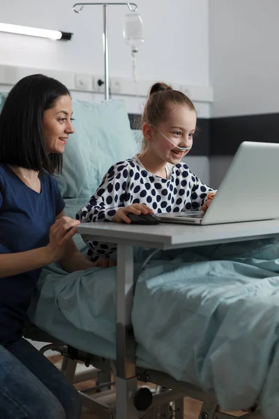 Happy sick little girl playing games on laptop while mother sitting beside her inside hospital pediatrics ward room. Joyful child under treatment gaming on patient bed while parent takes care of her.