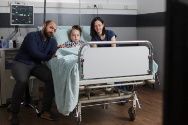 Considerate parents sitting next to hospitalized sick daughter while watching movies on television. Mother and father laying with ill little girl on patient bed while watching family film together.