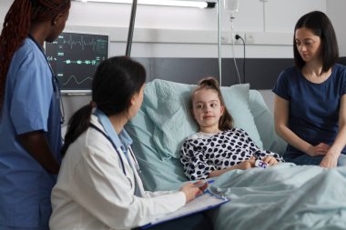 Pediatrist examining sick little girl disease symptoms in children healthcare facility room while mother sitting beside her. Ill daughter under treatment getting consulted by hospital medical staff.
