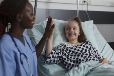 African american nurse doing high five gesture with sick girl resting in hospital pediatric ward patient bed. Childcare healthcare facility staff high fiving ill kid under medical treatment.