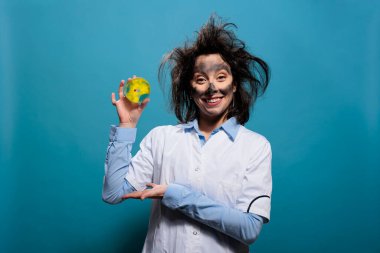 Mad chemist with dirty face and messy hair having petri dish glass plate with toxic biological material sample. Wacky microbiologist with experiment result on glass plate while on blue background.