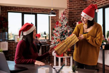 Asian woman receiving christmas present from coworker to celebrate xmas holiday in office with festive decorations and seasonal winter ornaments. Giving gifts to colleagues in business space.