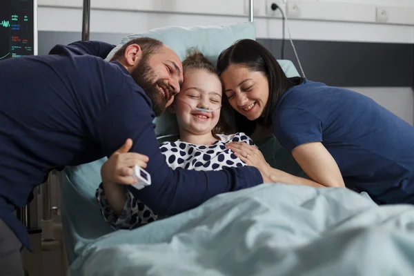Happy sick little girl hugged by joyful smiling parents in hospital pediatrics ward. Cheerful mother and father hugging ill daughter sitting in patient bed while under treatment.
