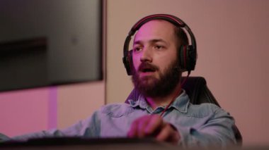 Handheld shot of man with gaming headset looking at computer screen and talking to team members while streaming gameplay. Gamer playing multiplayer online action game using professional pc setup.