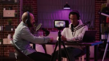 Static tripod shot of internet radio host recording podcast using digital video camera greeting guest in home studio. African american podcaster doing fist bump in interview with famous influencer.