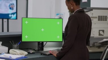 African american receptionist using greenscreen on computer, working at hospital reception desk. Looking at isolated mockup template with chroma key background and blank copyspace display.