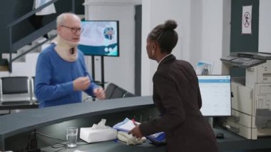 Medical worker helping injured patient with cervical collar at hospital reception counter, waiting to attend appointment with medic. Old man with neck brace in pain at health center.