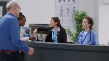 Medical assistant giving support to old patient at reception desk in hospital lobby, helping with checkup appointment and report papers. Nurse and senior man talking at registration counter.