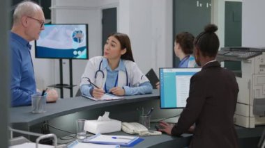 Asian doctor talking to senior patient at hospital reception desk, using report papers to fill in before attending medical appointment. Medic helping old man with consultation insurance.