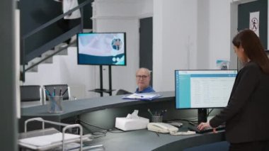 Senior man filling in checkup report papers before appointment, talking to asian receptionst at hospital registration counter. Patient with healthcare insurance at facility. Handheld shot.