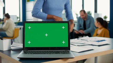 Laptop computer with green screen on desk in front of african american startup employee looking at paper with charts. Focus on portable computer screen with chroma key empty background.