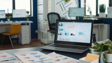 Closeup of laptop screen showing moving charts and financial data in empty modern office in startup company. Interior of marketing department with portable computer showing business data on desk.