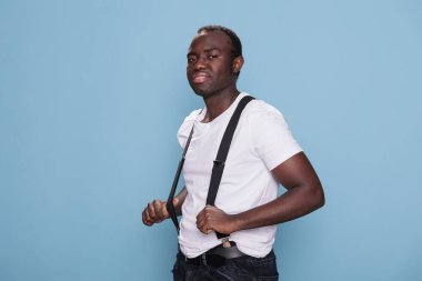 Happy young man smiling at camera while pulling suspenders. Portrait of confident cool looking african person standing on blue background wearing fashion clothes. Studio shot