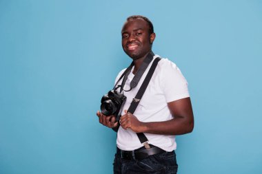 Confident professional photographer having modern camera while smiling heartily and standing on blue background. Joyful handsome looking african ethnicity guy with photo device posing with confidence.