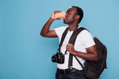 Tourist with camera sipping beverage while getting ready for urban journey. Photography enthusiast having DSLR device and backpack excited for holiday weekend voyage. Studio shot