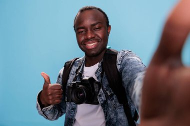 Confident photographer showing okay finger symbol while taking selfie and having DSLR camera on blue background. Photography enthusiast giving approve hand gesture while taking picture of himself.