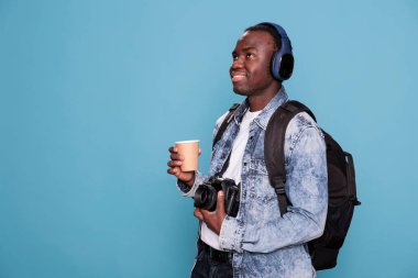 Smiling young man with professional camera and backpack going on holiday trip. Photography enthusiast having DSLR device and wireless headphones going on citybreak vacation.