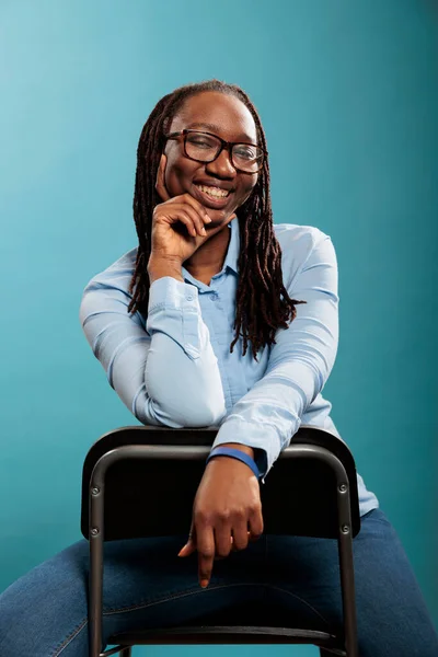 Excited cheerful african american pretty attractive woman sitting on chair while smiling at camera on blue background. Joyful pleased young adult person with confident smile and casual looking.