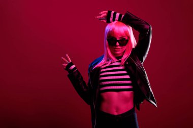 Carefree beauty model showing peace symbol with two fingers, wearing trendy sunglasses and stylish leather jacket. Modern attractive woman feeling sensual in fluorescent light.