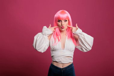 Confident beauty model with pink hair showing kissy face on camera, posing with carefree attitude in studio. Attractive young adult with trendy stylish makeup feeling sensual and pretty.